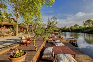 A wooden deck with outdoor seating, a view of a river, and a building with a tiled roof in the background.