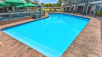 Piscina com deck de tijolos, cercada por uma grade e um edifício com sacadas.