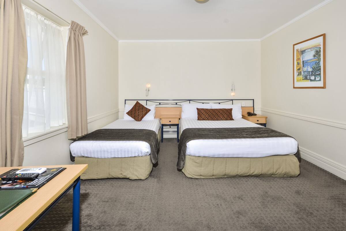 Habitación de hotel con dos camas individuales, un escritorio con silla, una mesita de noche y una ventana con cortinas.