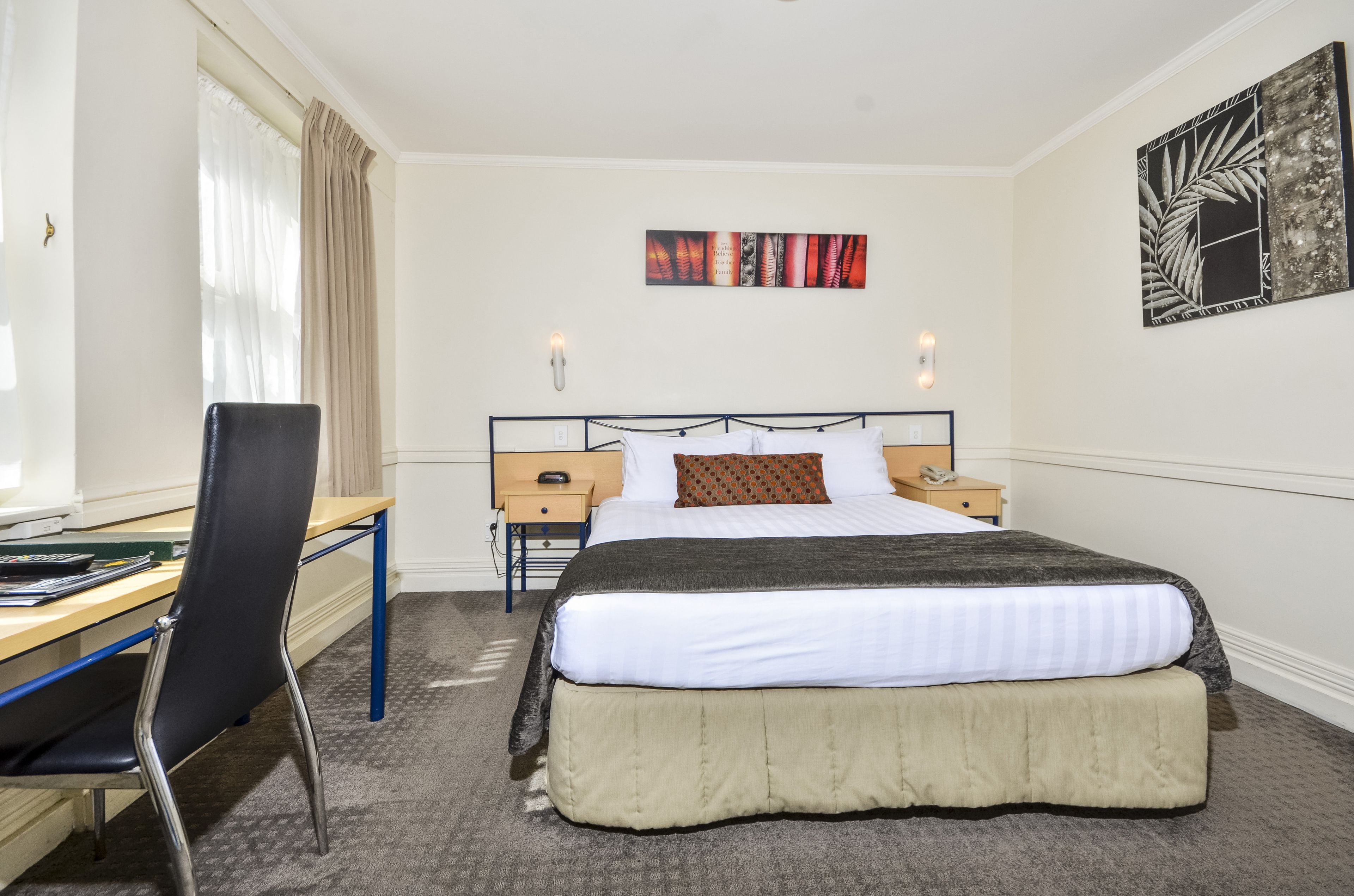 A hotel room with a bed, a desk, a chair, a nightstand, and wall art.