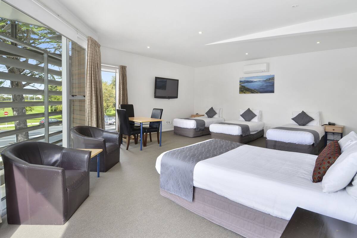 Habitación de hotel con una cama grande, dos camas más pequeñas, un escritorio y un sofá. Hay un televisor en la pared y una ventana con vistas al exterior.