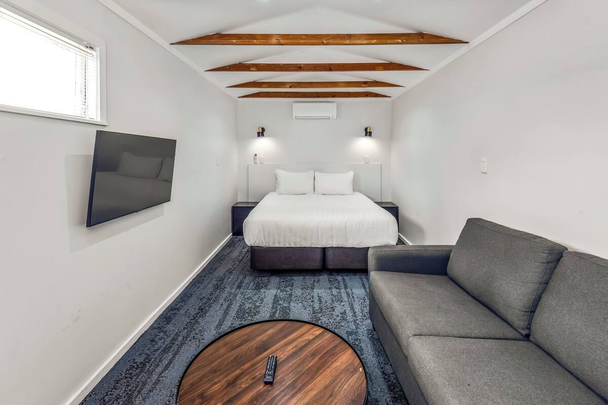 Una habitación compacta con una cama, un sofá gris, una mesa de centro redonda de madera y un televisor montado.