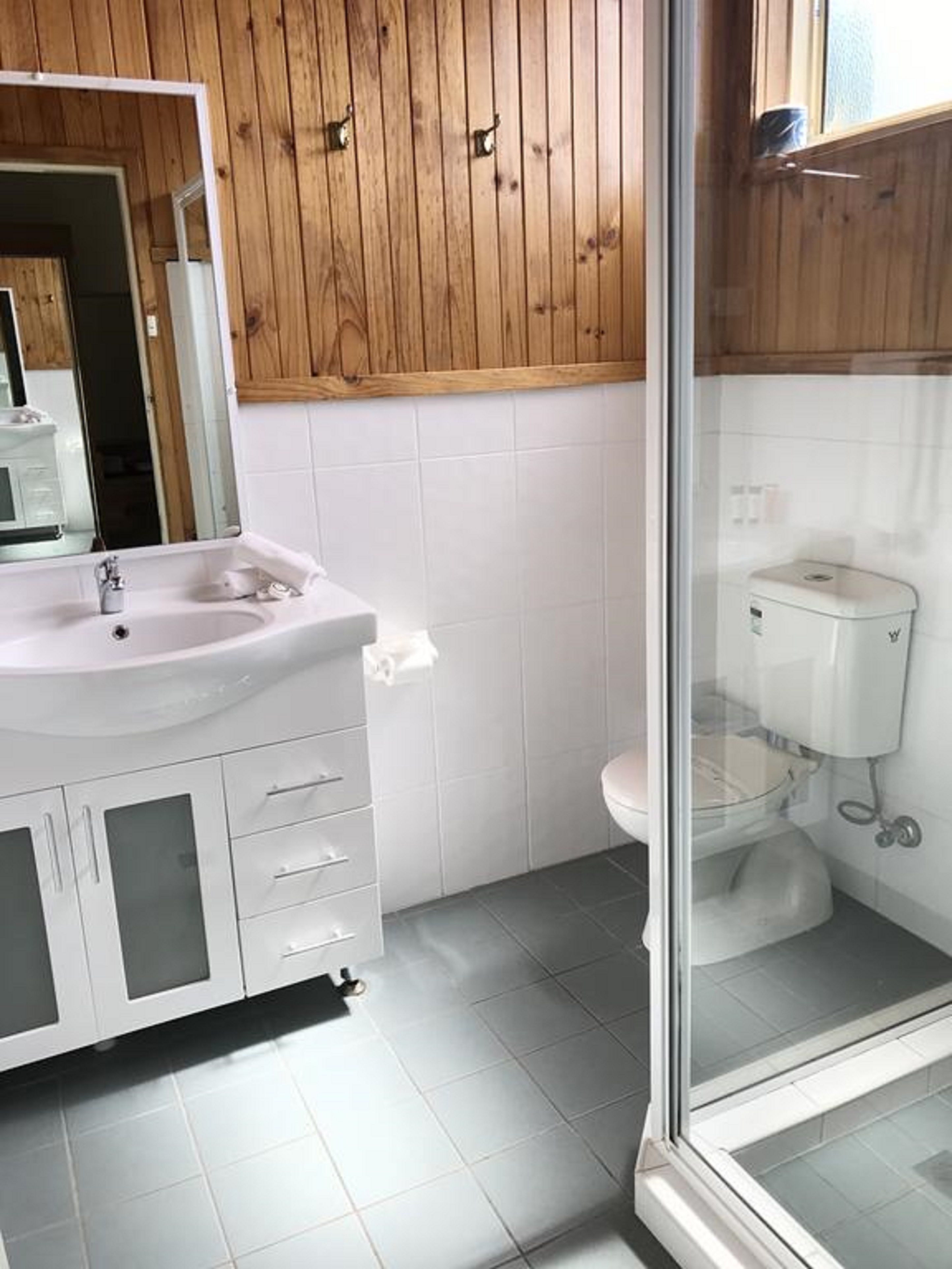 A bathroom with a white sink, mirror, and toilet. There is a wooden panel wall above the sink and a glass shower door.