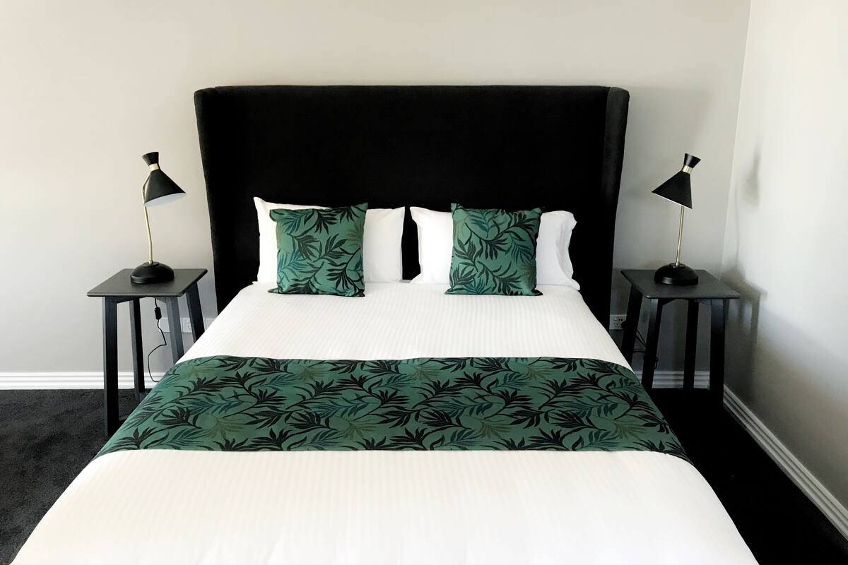 A bed with a black headboard, white bedding, and green leaf-patterned pillows. There are two black nightstands with lamps on either side.