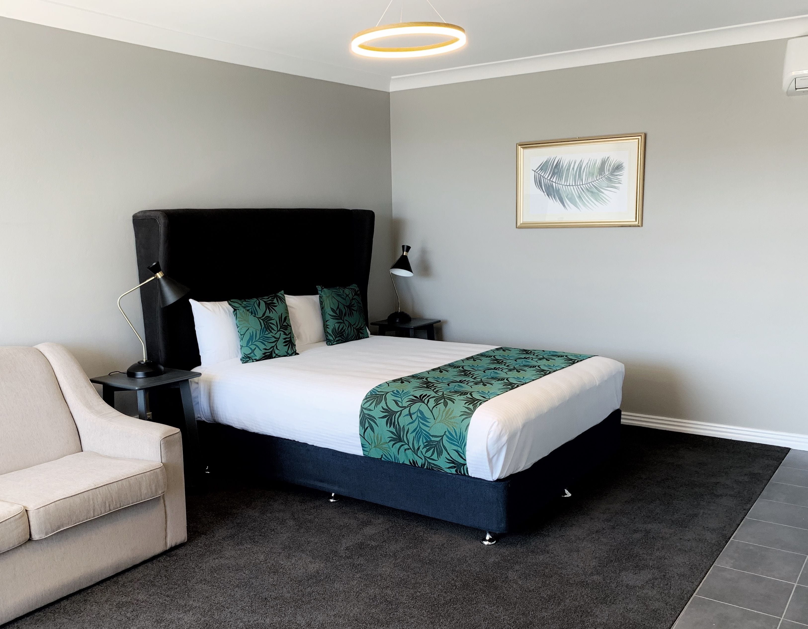 Un dormitorio ordenado con cama, mesita de noche, lámpara, silla y una obra de arte enmarcada en la pared.