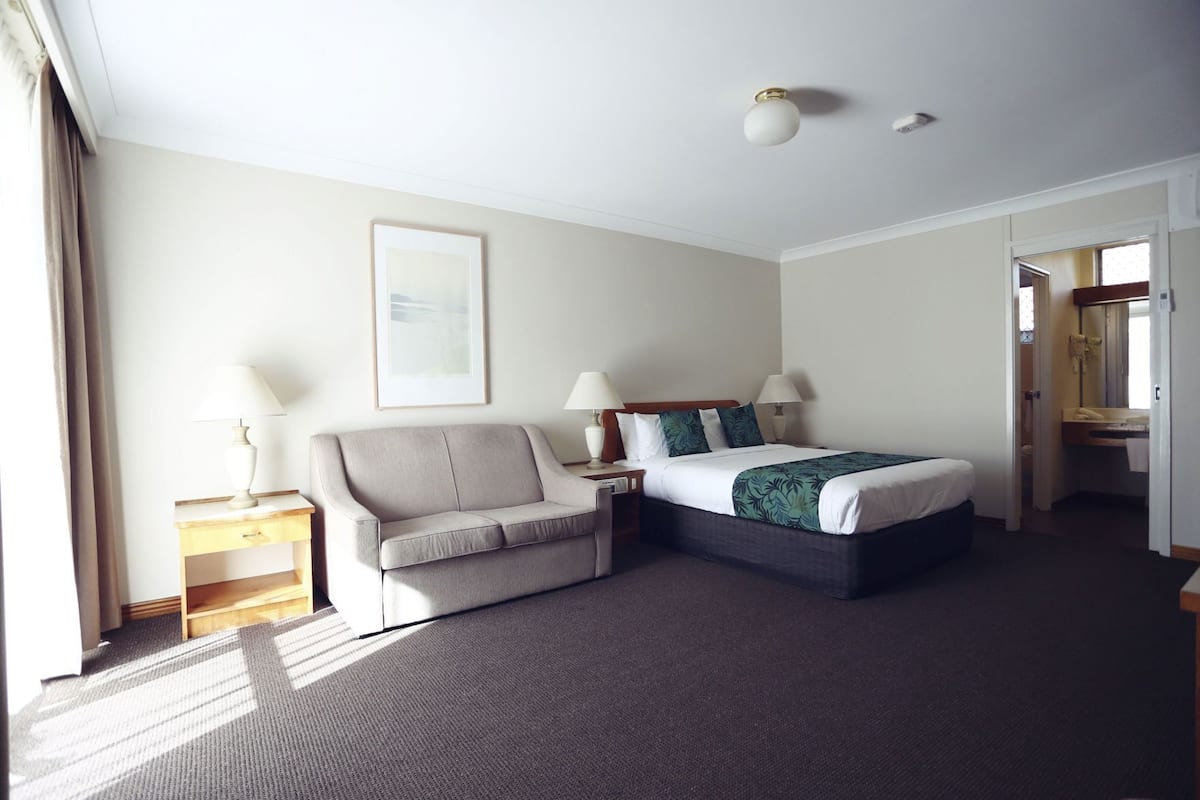 A hotel room with a bed, a sofa, a bedside table, a lamp, and a framed picture on the wall.