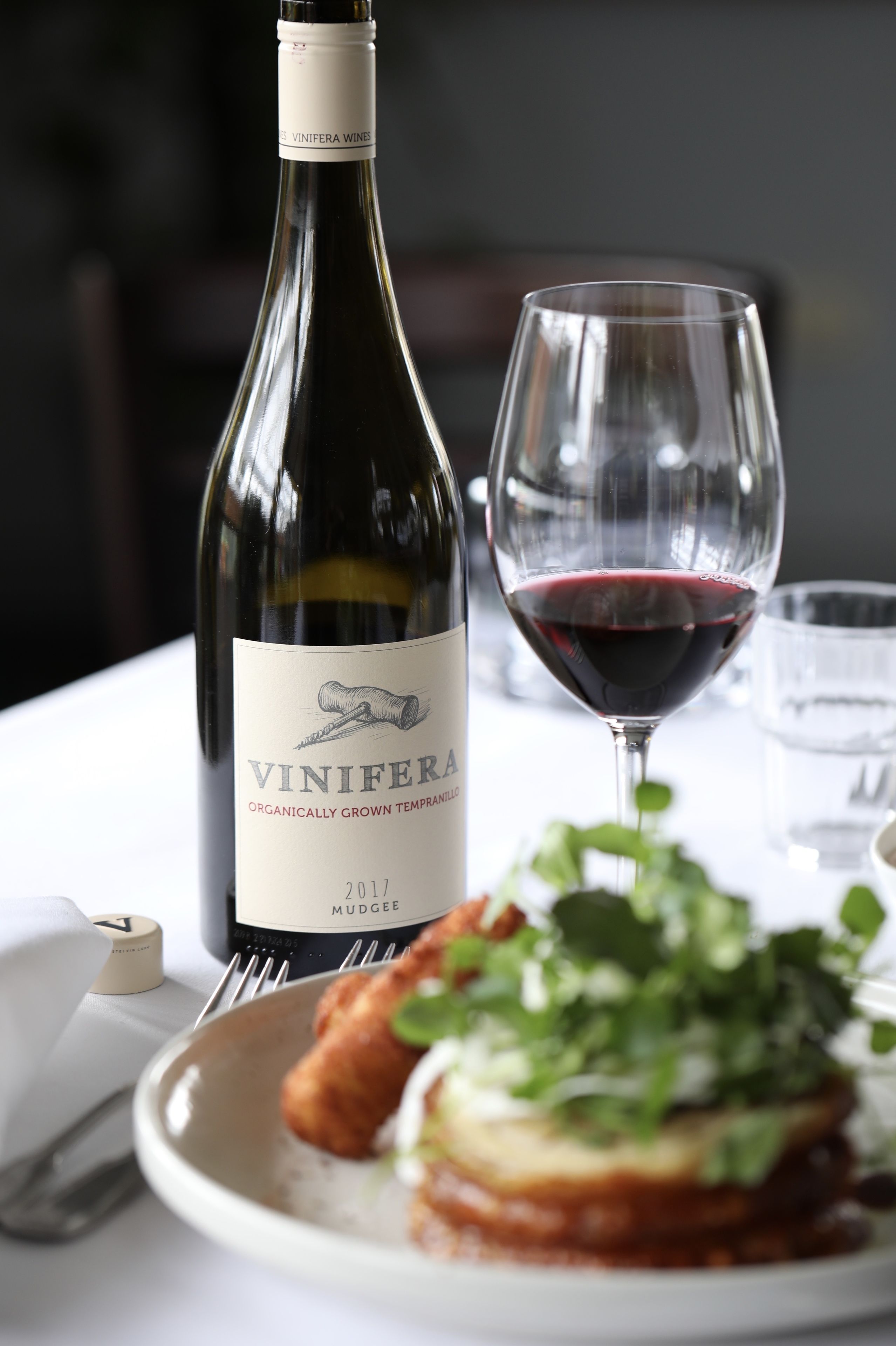 A bottle of Vinifera wine, a glass of red wine, and a plate with food on a table.