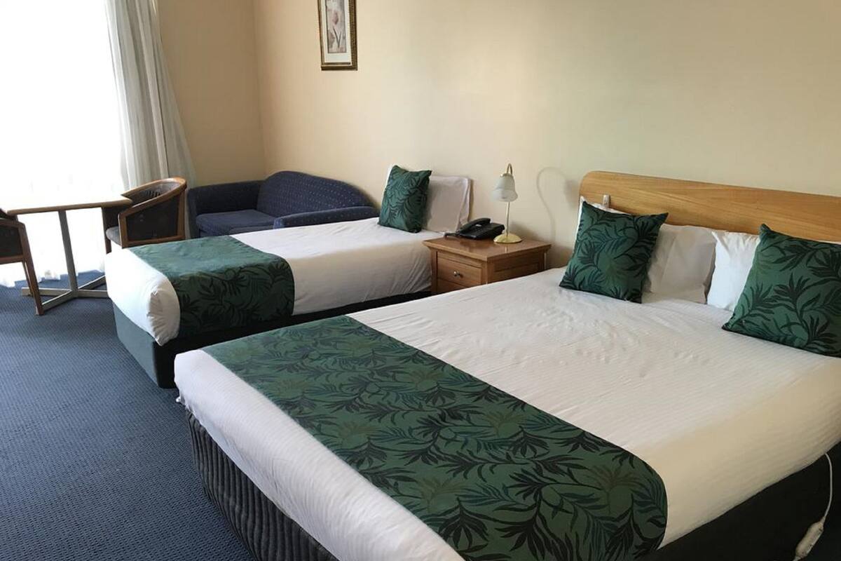 A hotel room with two beds, each with green leaf-patterned bedspreads, a wooden nightstand with a lamp, and a small table with a chair.