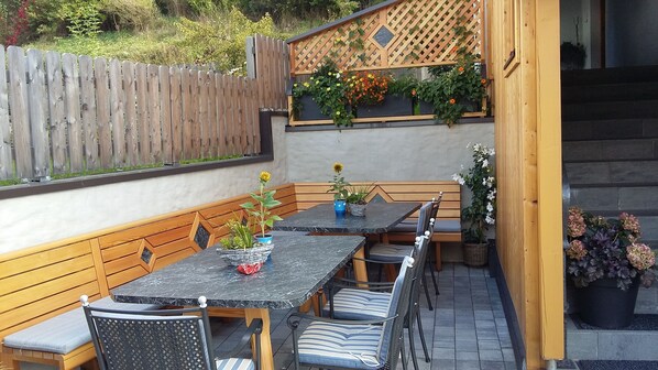 Un patio exterior con un banco de madera, una mesa y sillas. Hay plantas en macetas y una valla de madera al fondo.