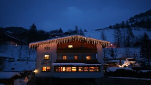 Een gebouw met twee verdiepingen, een met sneeuw bedekte kap en verlichte ramen, omgeven door een besneeuwd landschap en bomen.