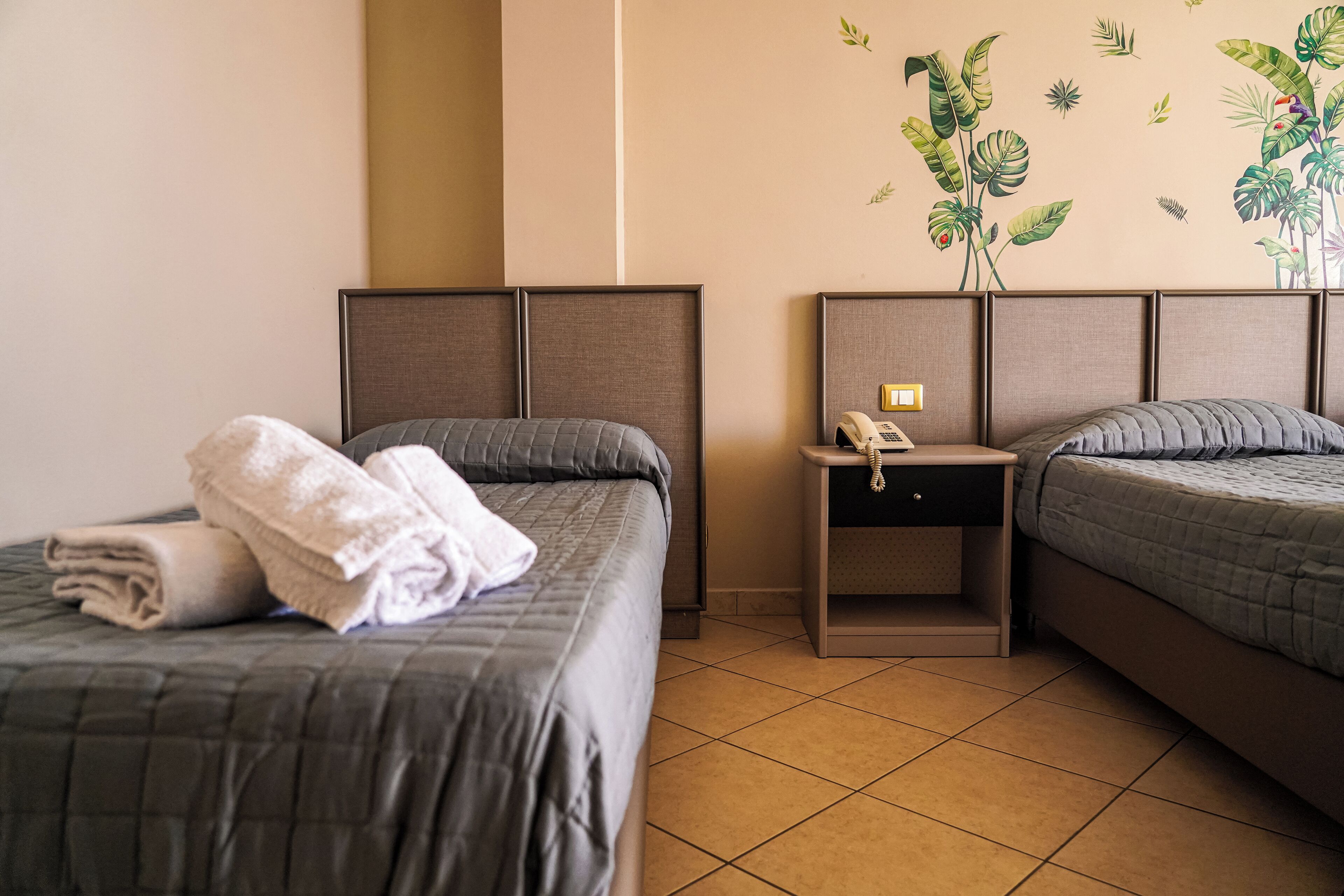 A hotel room with two beds, a nightstand with a phone, and a wall with a tropical plant decal.
