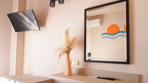 Una habitación de hotel moderna con un televisor de pantalla plana montado en la pared, un jarrón con plumas de pampa y una obra de arte enmarcada que representa un sol y olas.