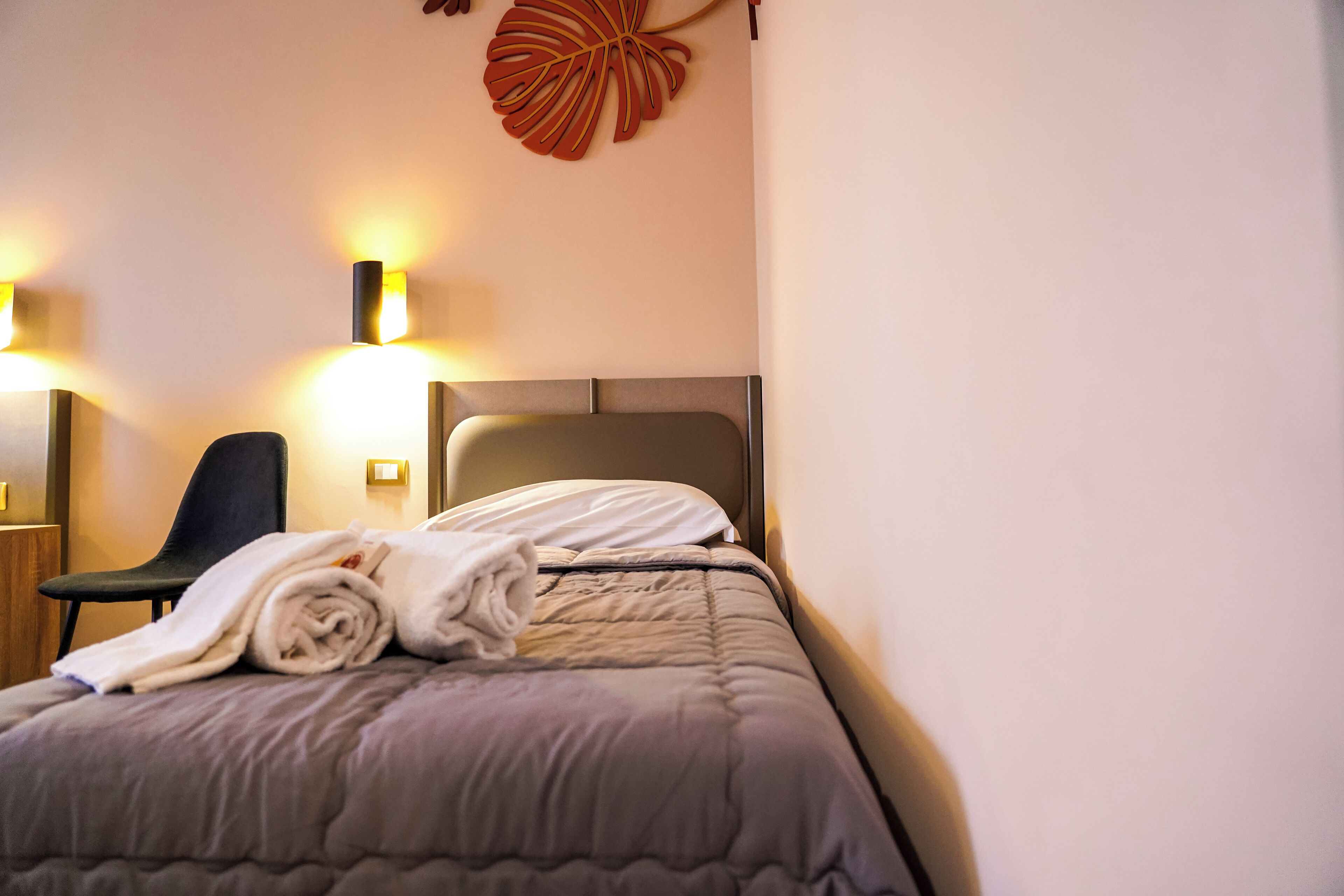 A neatly made bed with a grey duvet, a rolled white towel, and a chair to the left.
