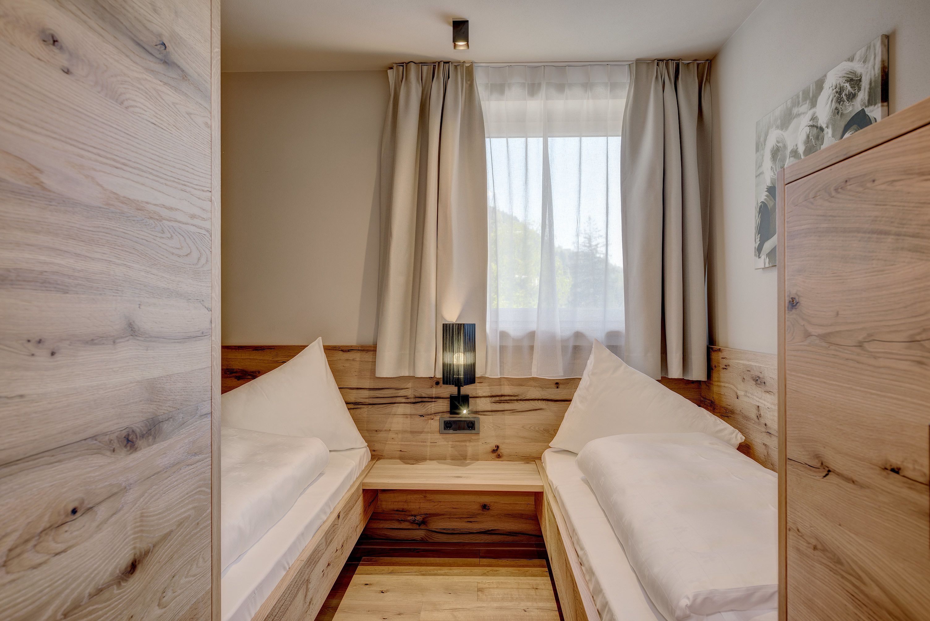A room with two beds, wooden walls, a window with sheer curtains, and a wall-mounted lamp.