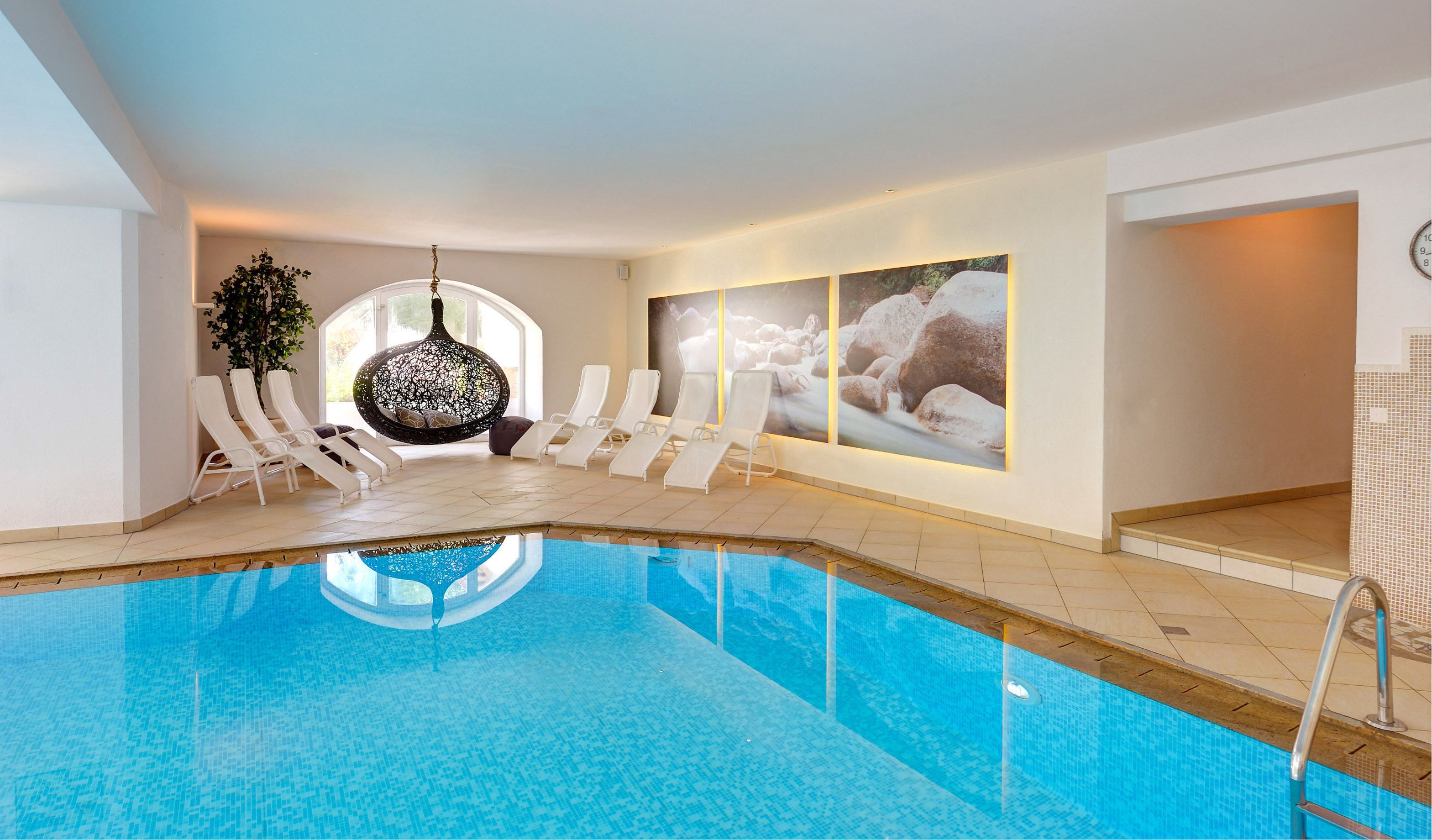 An indoor swimming pool with white lounge chairs and a hanging chair, surrounded by large framed pictures and a potted plant.