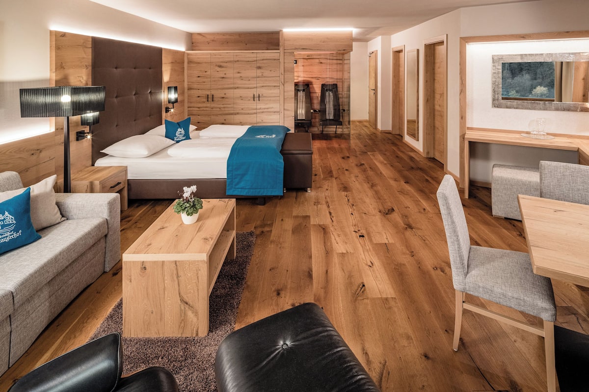 A modern hotel room with a bed, sofa, coffee table, and a dining area with chairs and a table.