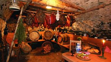 A rustic cellar with wooden barrels, hanging meats, and a wooden table with a candle, wine bottles, and a basket of apples.