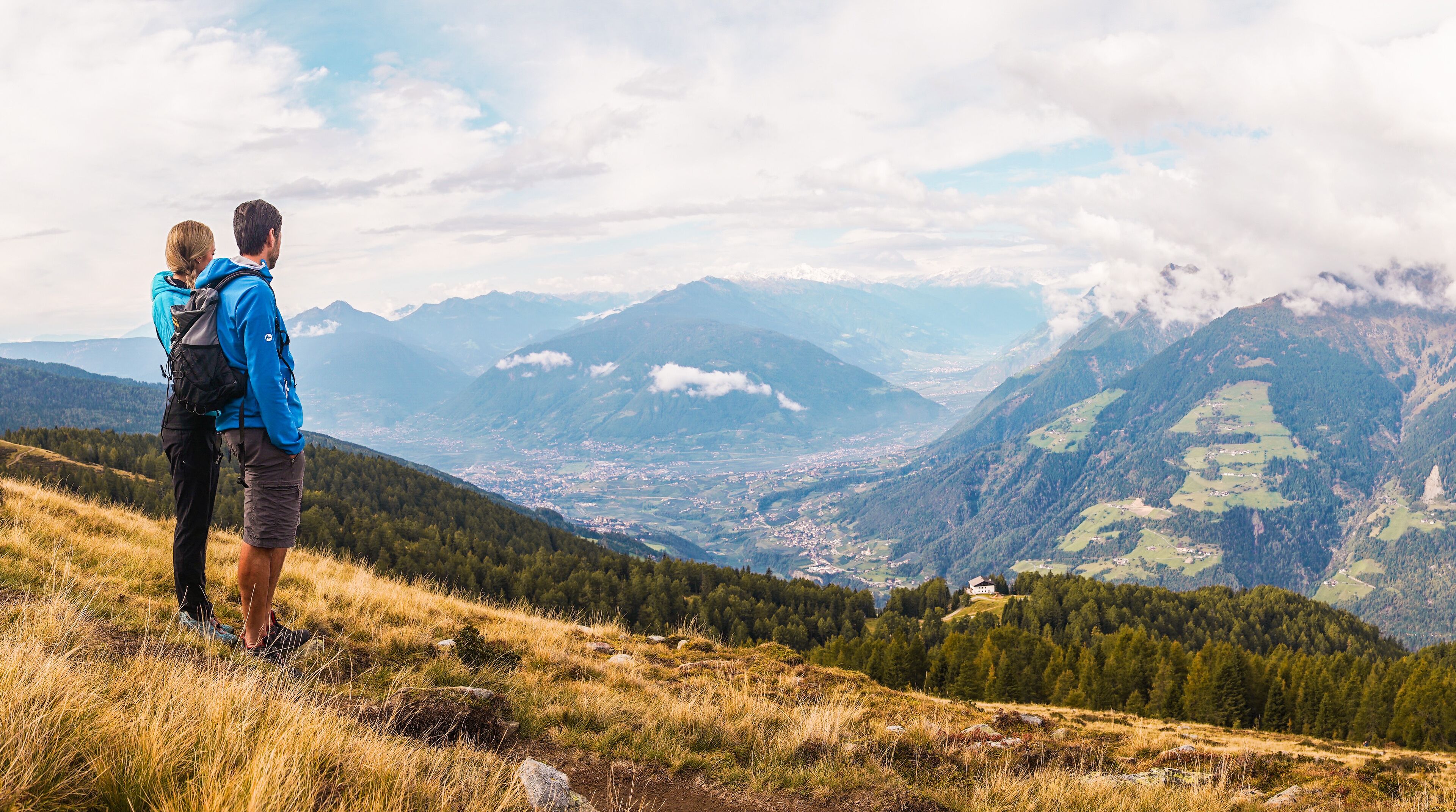 Un couple se tient sur une colline herbeuse, d’où l’on aperçoit une vallée et, au loin, des montagnes.