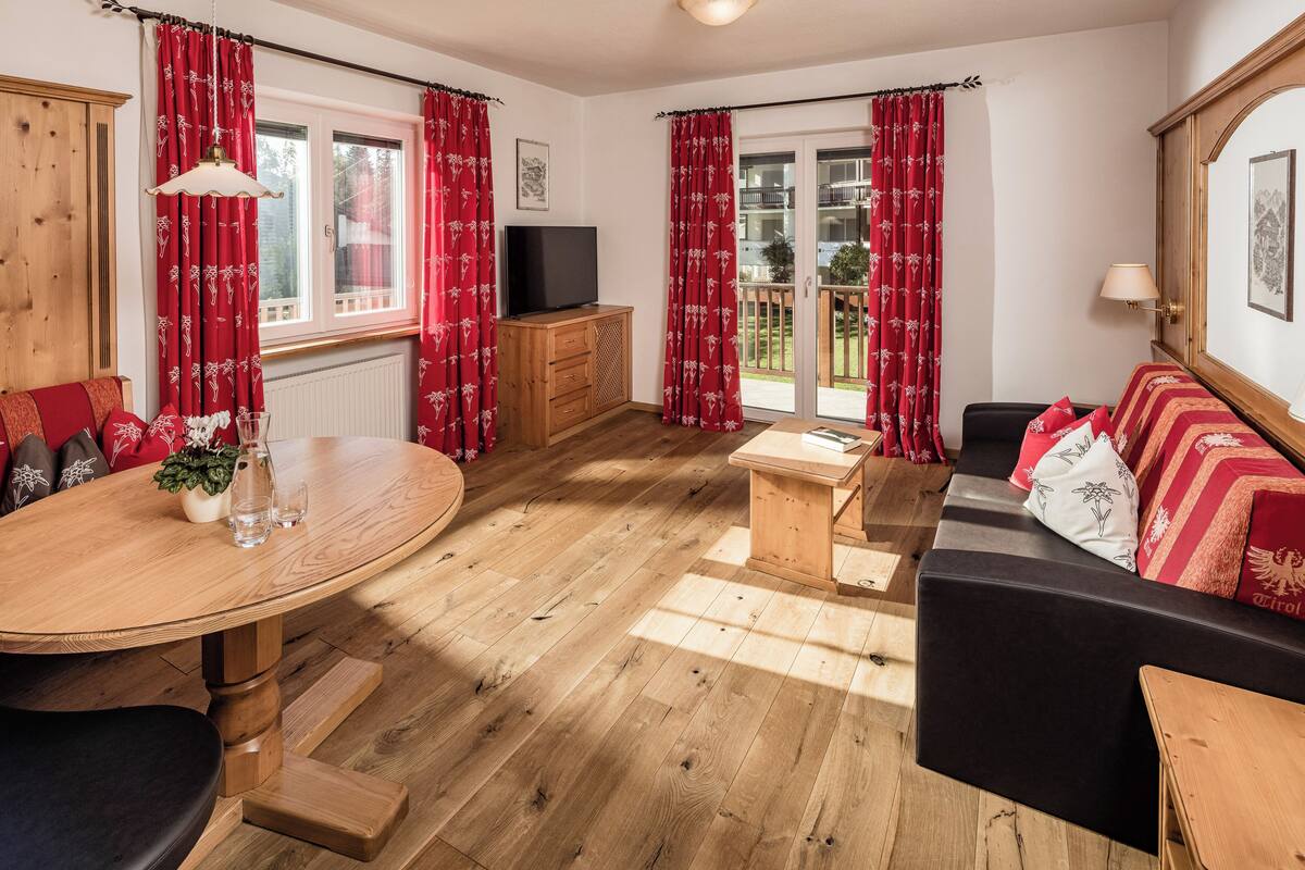 A room with a wooden floor, a sofa, a TV, a wooden cabinet, a dining table, and red curtains.