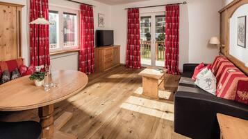 A room with a wooden floor, a sofa, a TV, a wooden cabinet, a dining table, and red curtains.
