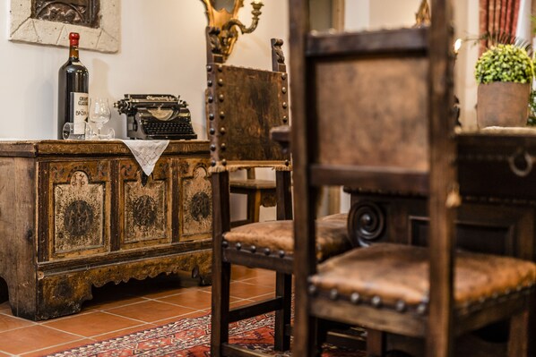 A vintage wooden chair with a cushioned seat and backrest, positioned in front of a wooden cabinet with a wine bottle and glasses on top.