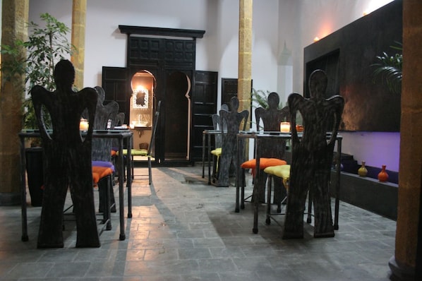 A dimly lit interior with a central seating area featuring chairs with cushions, a fireplace, and a wall-mounted sculpture. - Riad 7 (Azemmour)