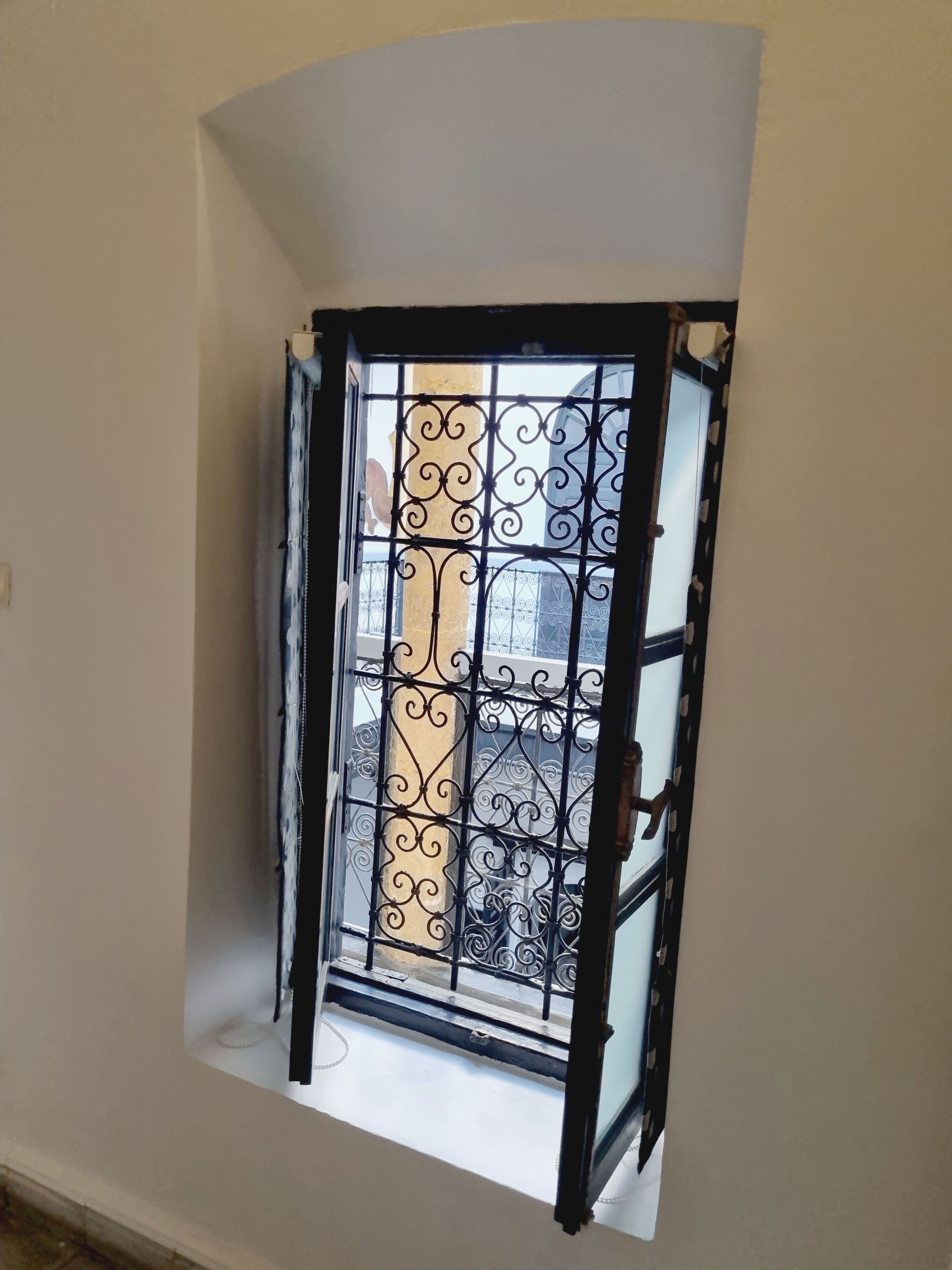 A room with a curved wall, a window with decorative iron bars, and a view of buildings outside.