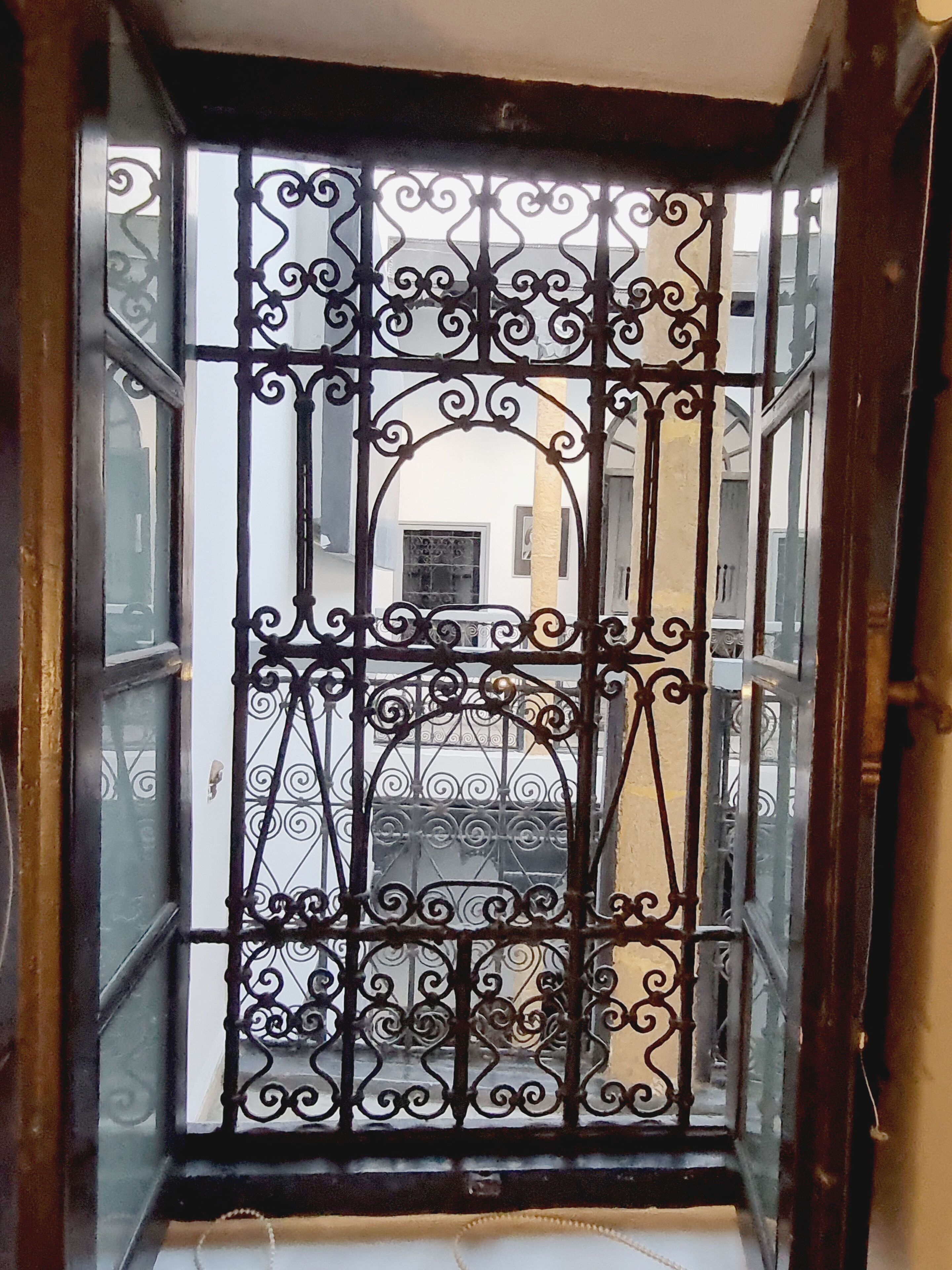 A view through a barred window with a balcony, featuring a building with a yellow facade and a white window.