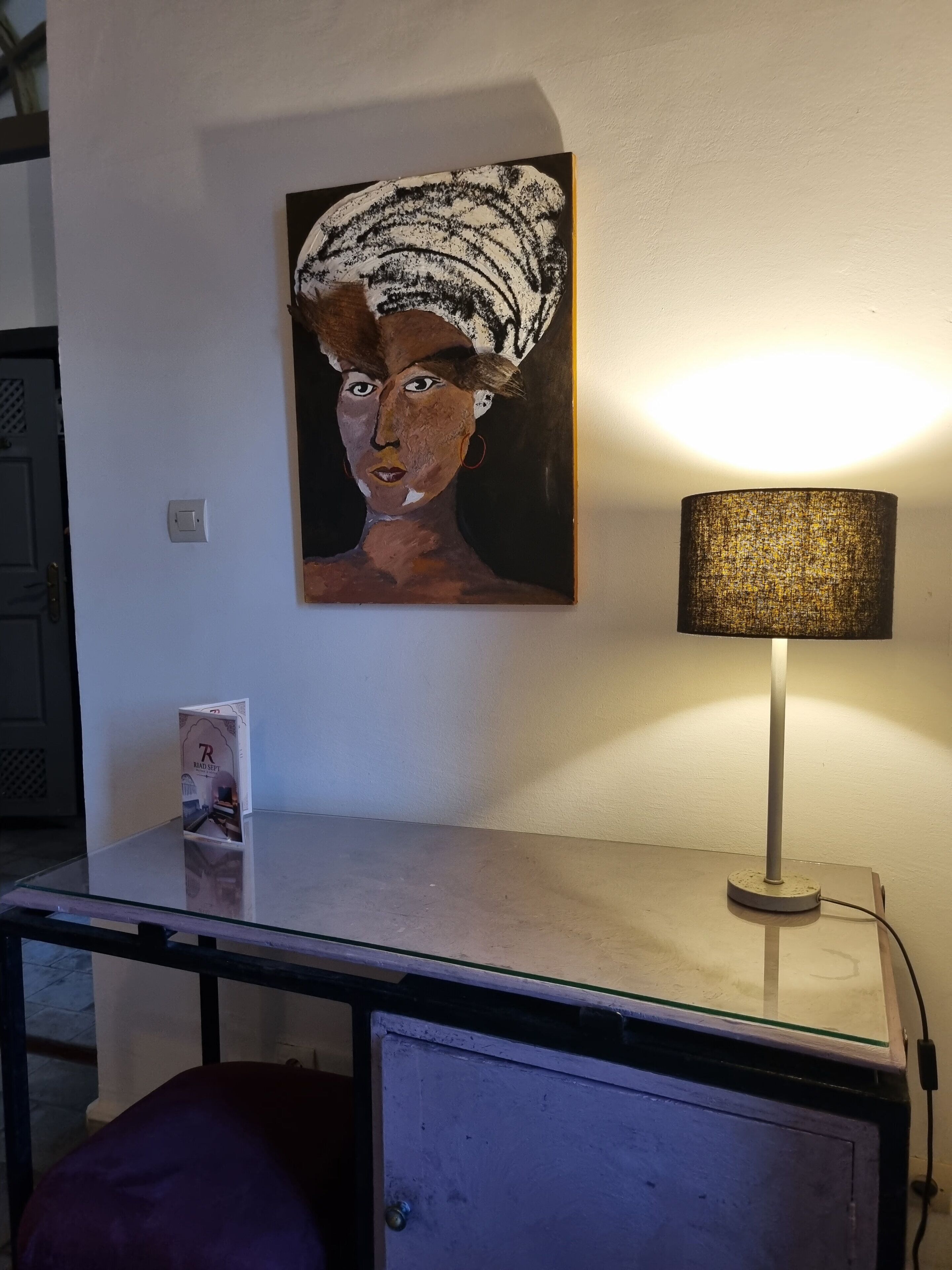 A desk with a lamp, a painting, and a small card on the wall.