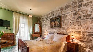 A bedroom with a stone wall, a bed with white linens, a wooden nightstand, a bedside table, a mirror, and a painting. - Hotel Monte Cristo (Kotor)