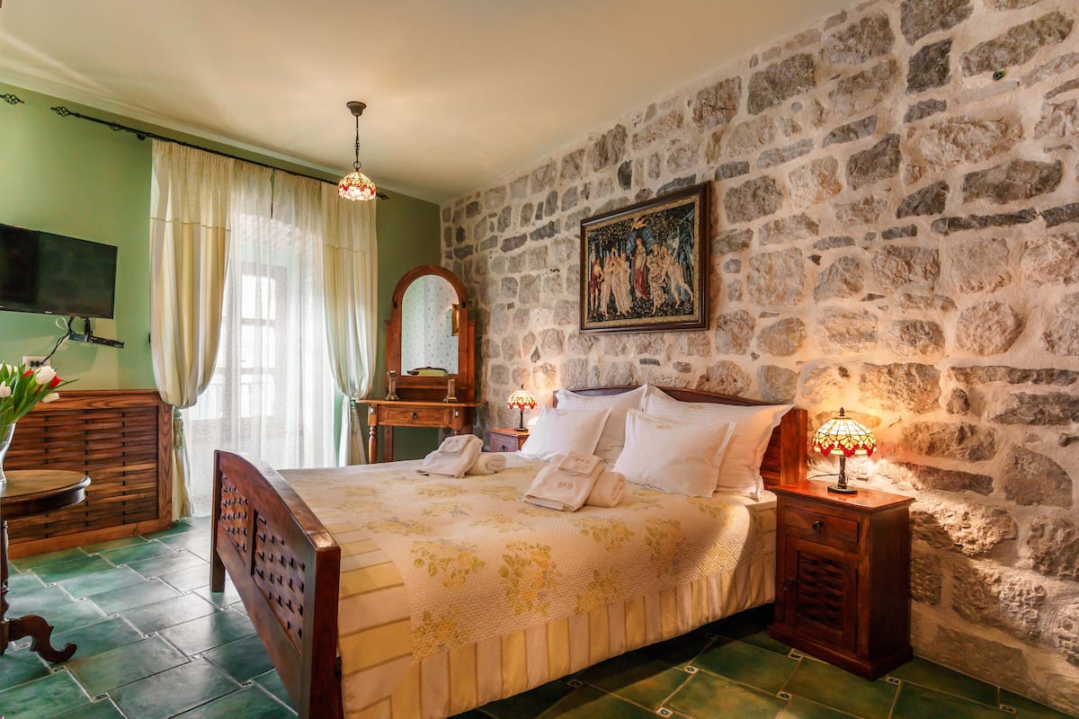 A bedroom with a stone wall, a bed with white linens, a wooden nightstand, a bedside table, a mirror, and a painting.