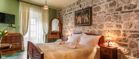 Un dormitorio con pared de piedra, una cama con sábanas blancas, una mesita de noche de madera, una mesita auxiliar, un espejo y un cuadro.