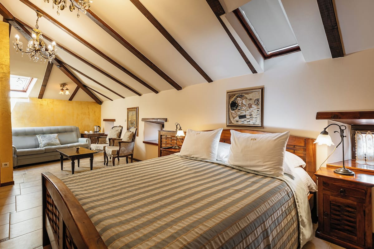 A spacious bedroom with a large bed, wooden ceiling beams, a chandelier, a sofa, and a desk with a lamp.