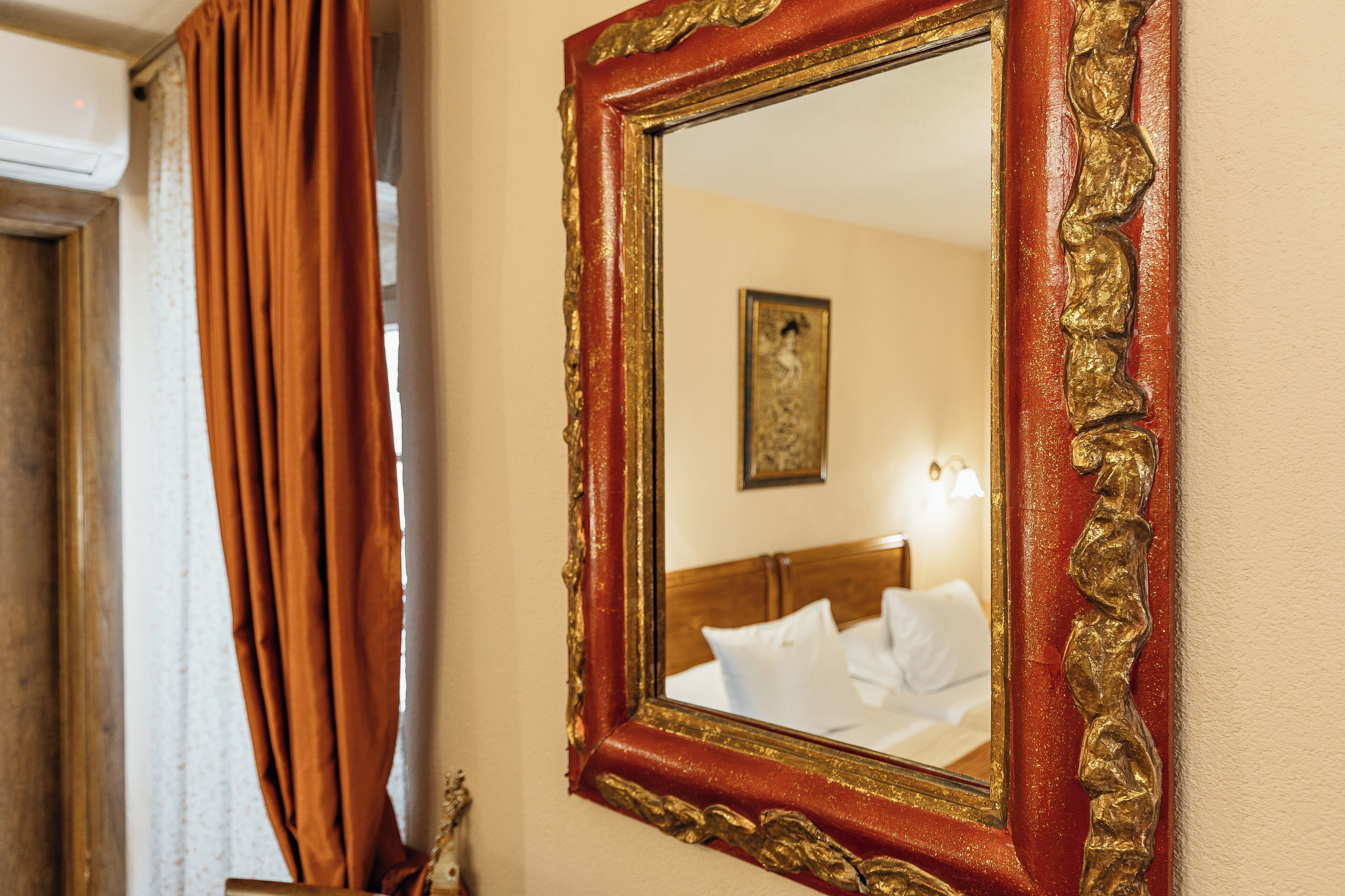 A mirror with a red frame and gold detailing, reflecting a bed with white bedding and a painting on the wall.