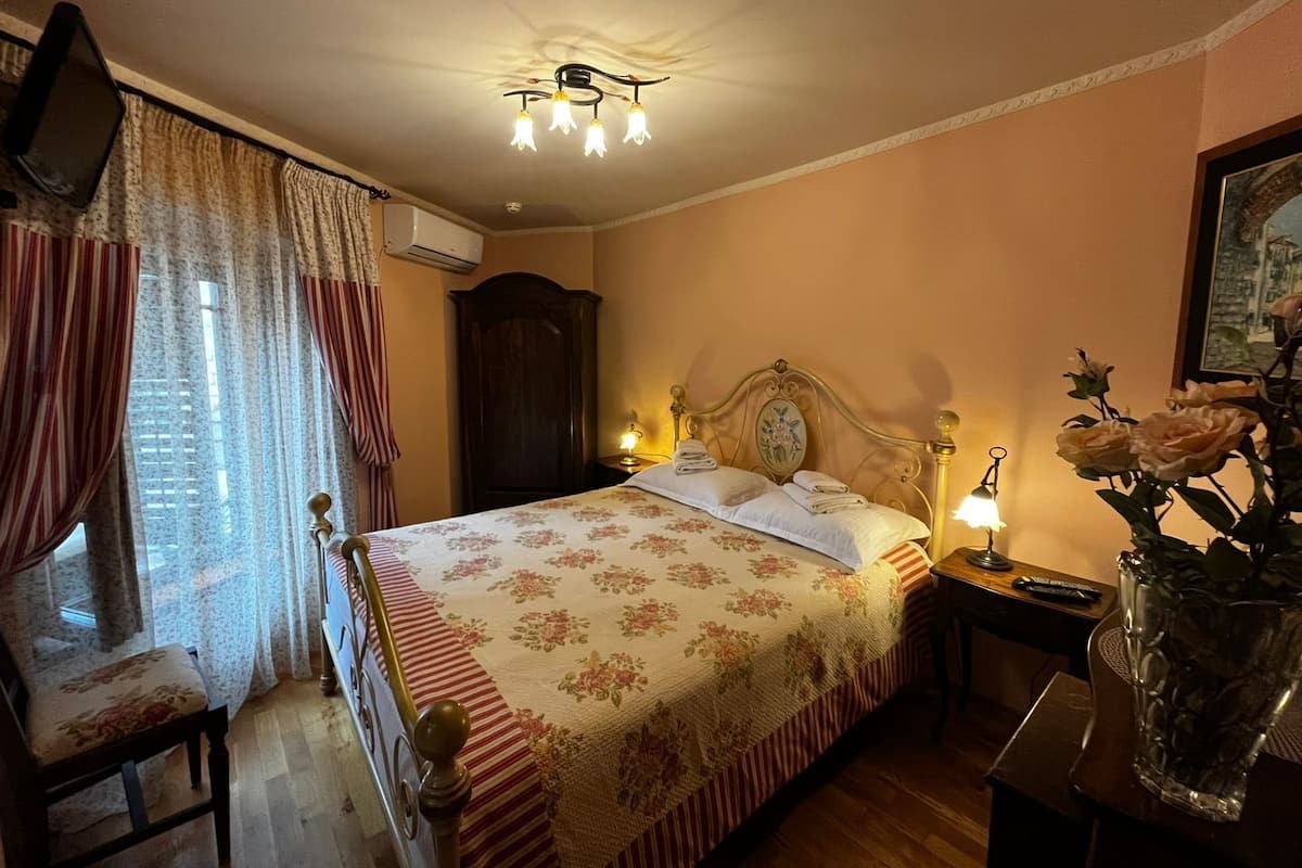 A bedroom with a bed, curtains, a nightstand, a lamp, a vase with flowers, and a painting on the wall.