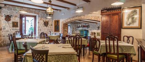 A rustic dining area with stone walls, wooden beams, and traditional decor. Tables are set with green tablecloths and wooden chairs.