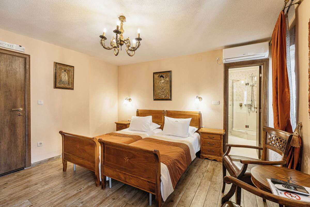A bedroom with a wooden bed, bedside tables, a chandelier, and a bathroom visible through an open door.