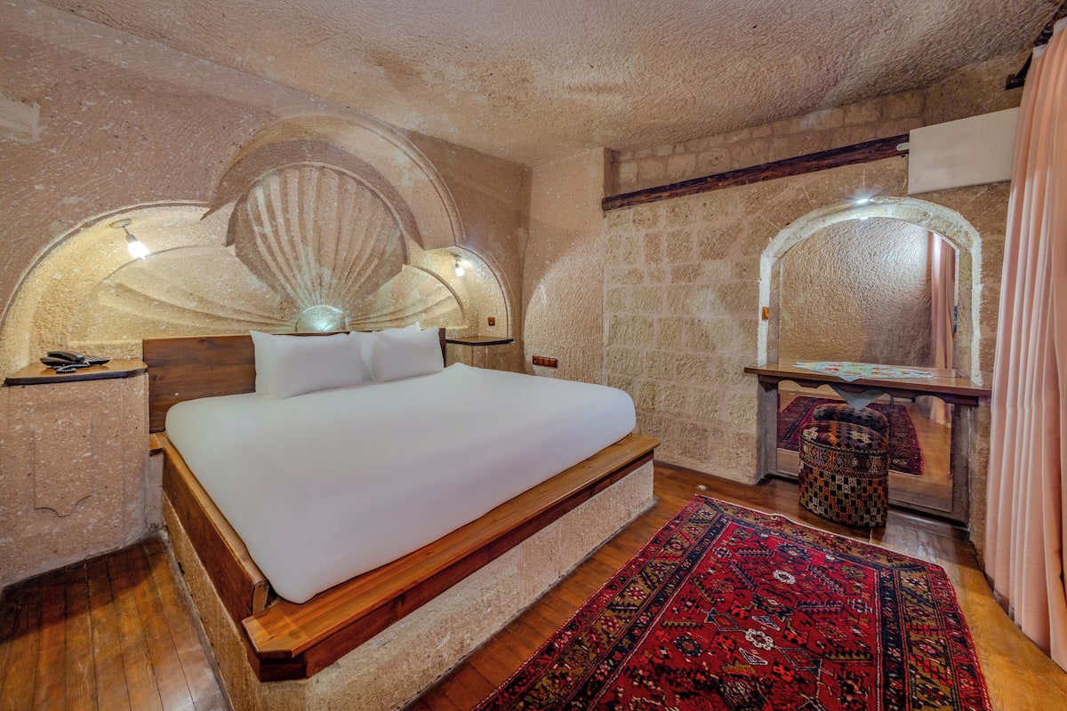 A bedroom with a large bed, a wooden headboard, a stone wall, a mirror, and a red patterned rug.