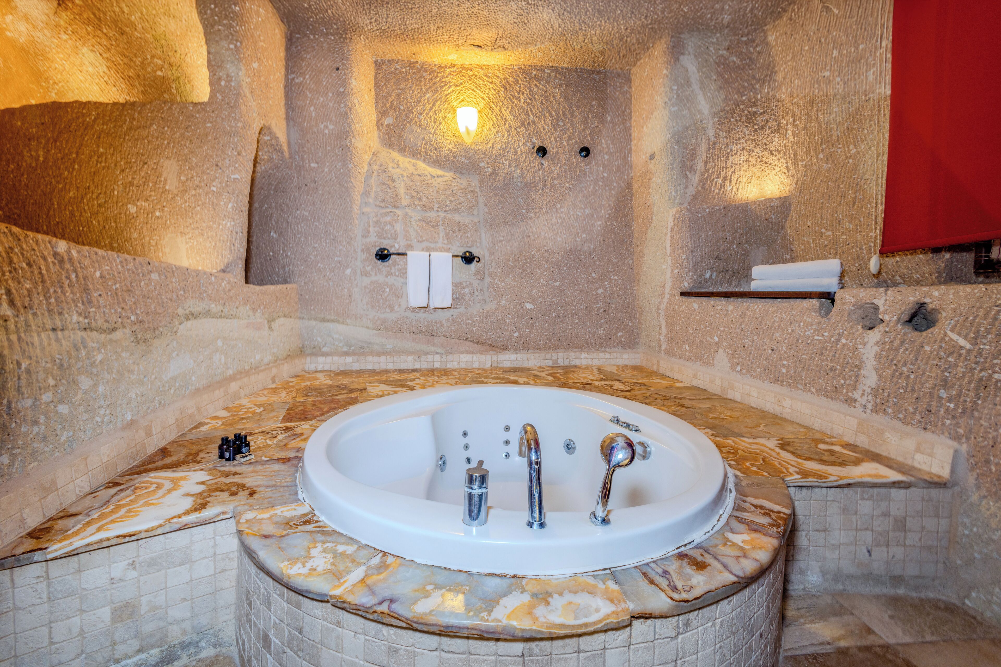 A stone-walled bathroom with a marble-topped bathtub, a towel rack, and a shelf with toiletries.