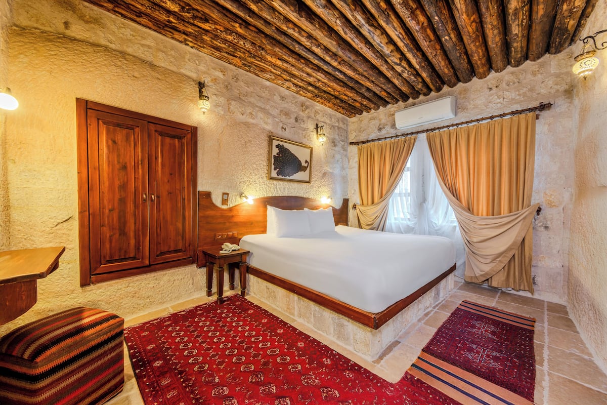 A room with a wooden ceiling, a bed with white linens, a red patterned rug, and a wooden wardrobe.