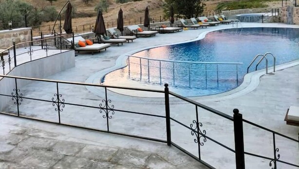 Un área de piscina con sillas de descanso y sombrillas, rodeada por una terraza de piedra y una valla de metal.