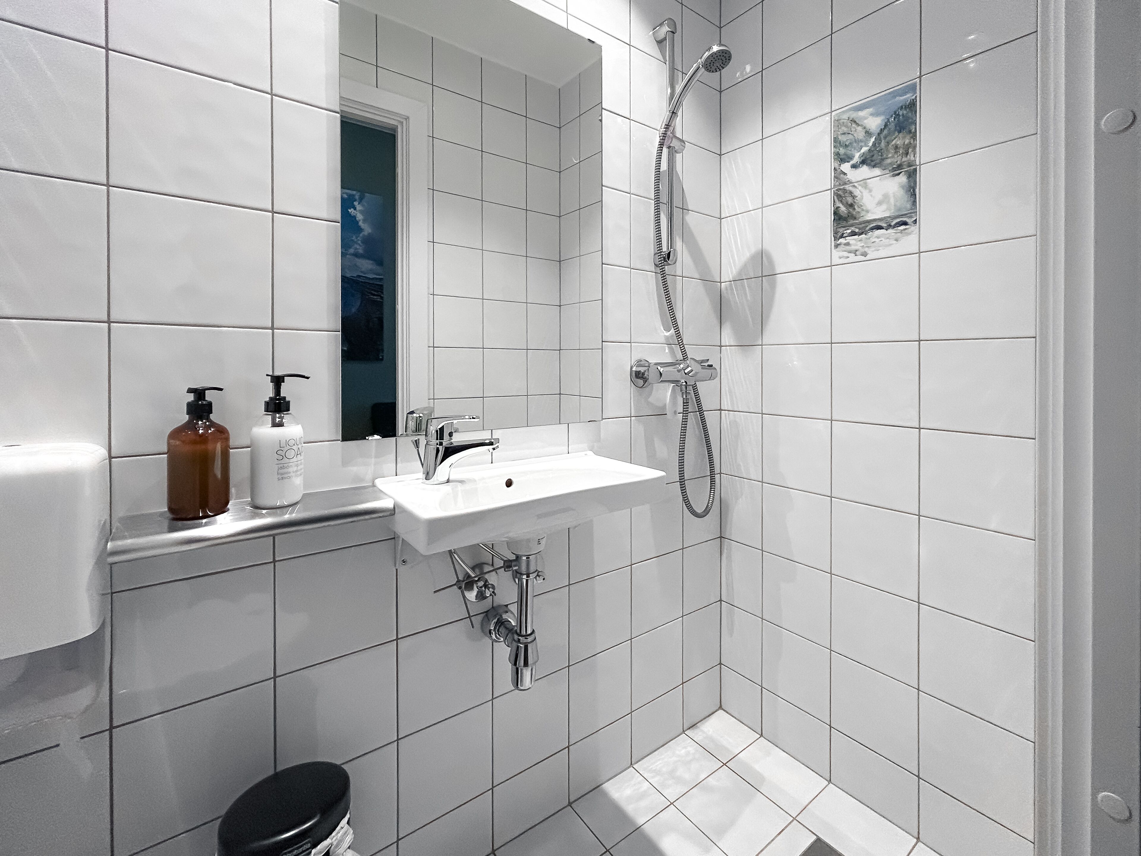 Een moderne badkamer met witte tegels, een wastafel, een spiegel, een douche en twee flessen zeep.