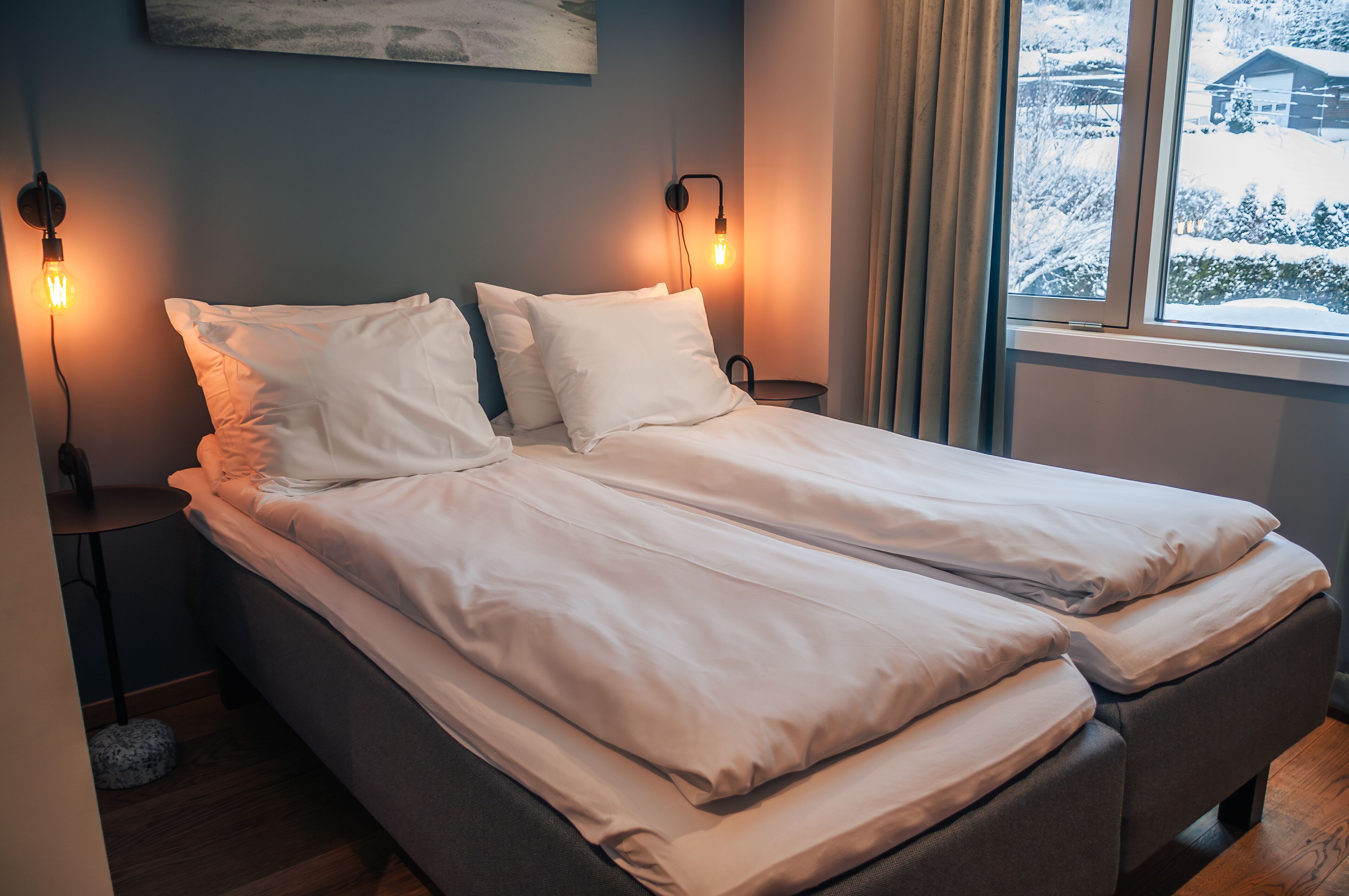 A neatly made bed with white linens, two bedside lamps, a small round table, and a window with a snowy view.