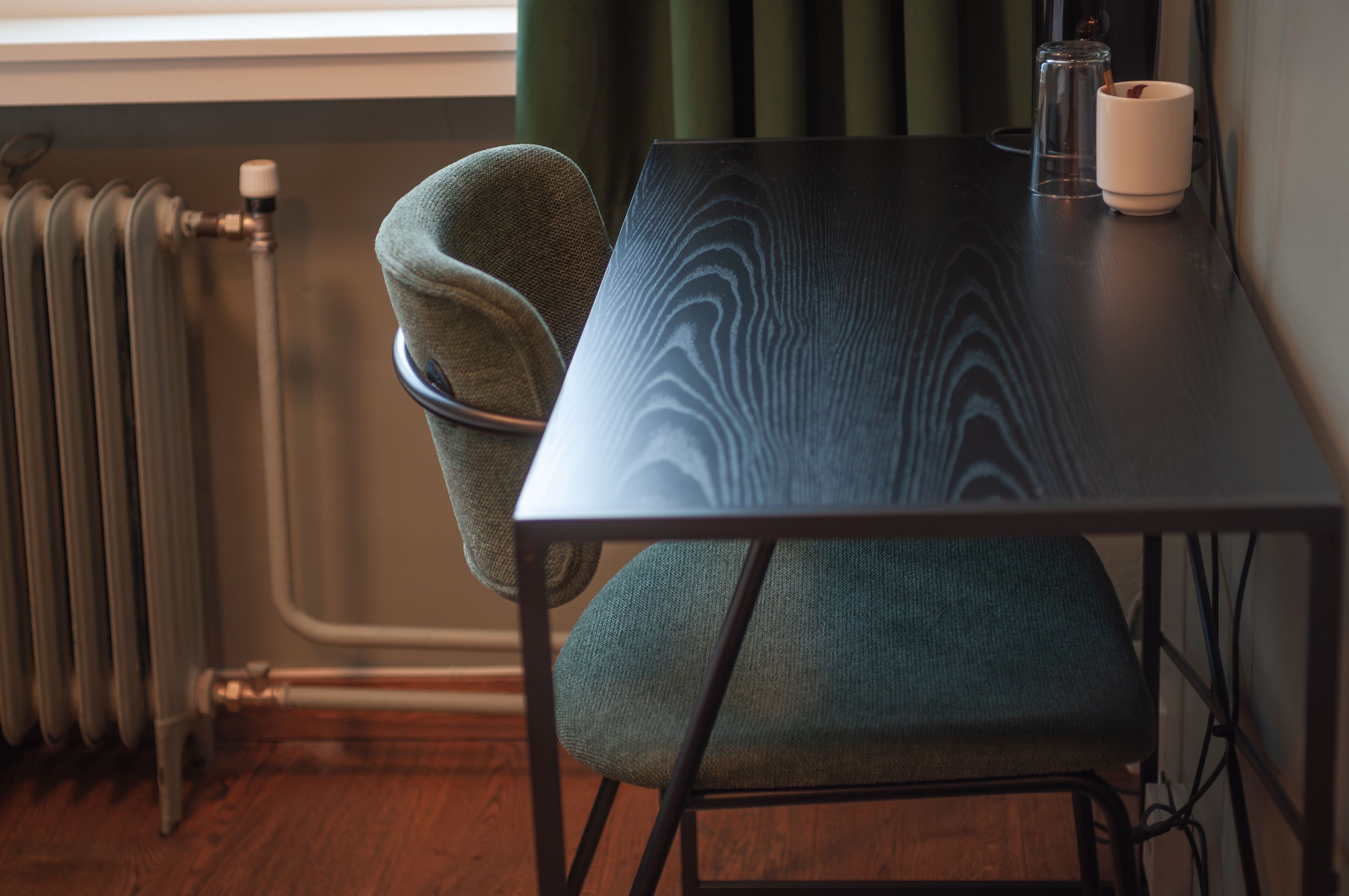 A room with a wooden floor, a radiator, a table with a chair, and a window with curtains.
