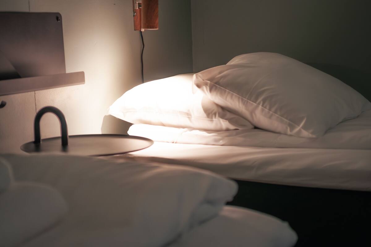 A bed with white bedding and a black headboard, a bedside table with a lamp, and a wall-mounted power outlet.