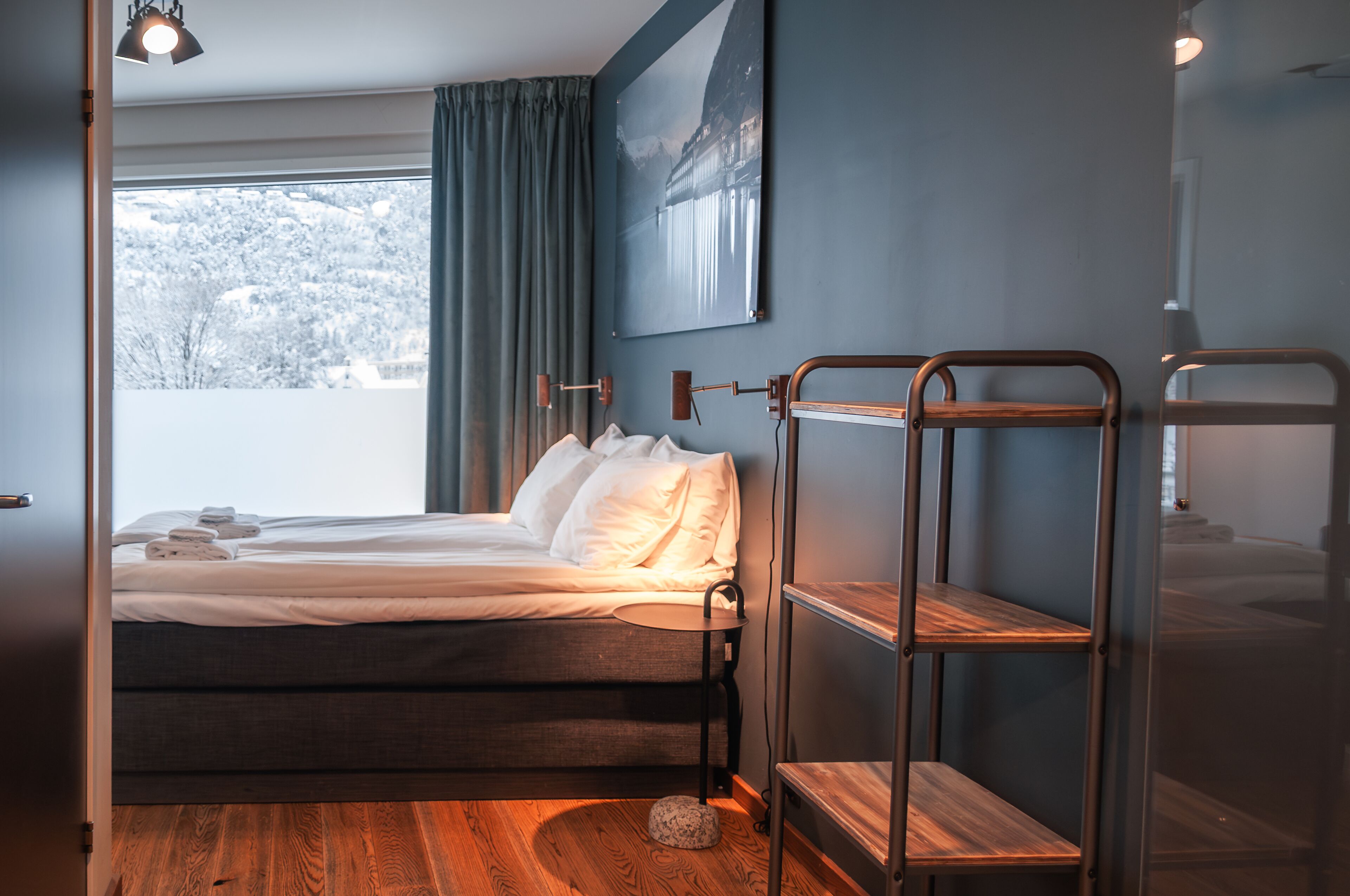 A bedroom with a bed, a window with curtains, a wall-mounted shelf, and a painting.