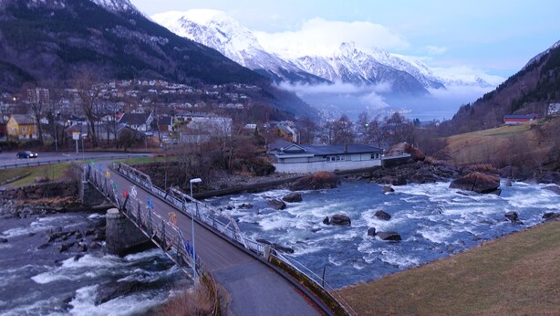 A bridge over a fast-flowing river with a town and mountains in the background. - Trolltunga Hotel (Ullensvang)