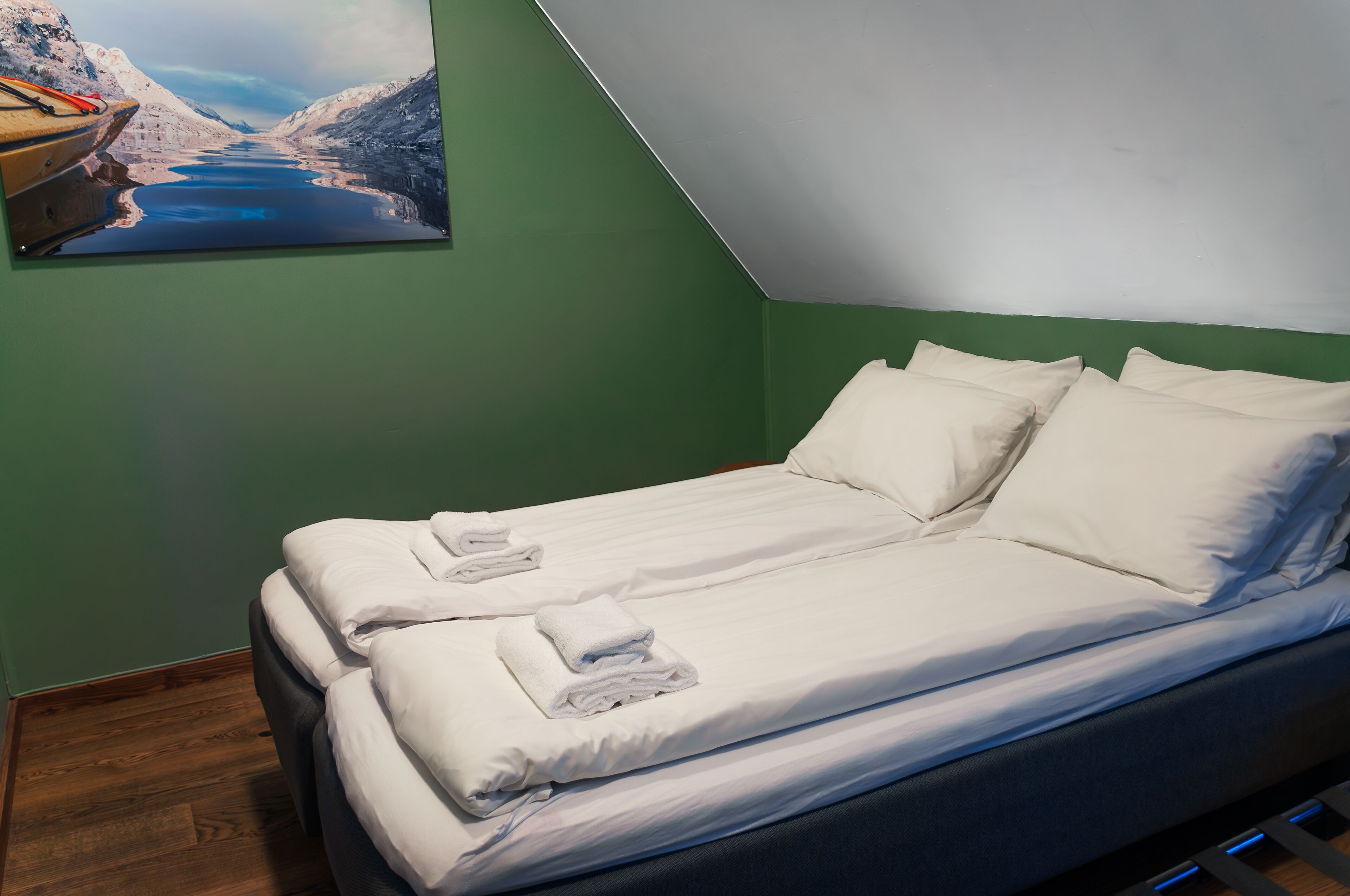 A neatly made bed with white linens and pillows, a painting of a mountainous landscape on the wall, and a wooden floor.