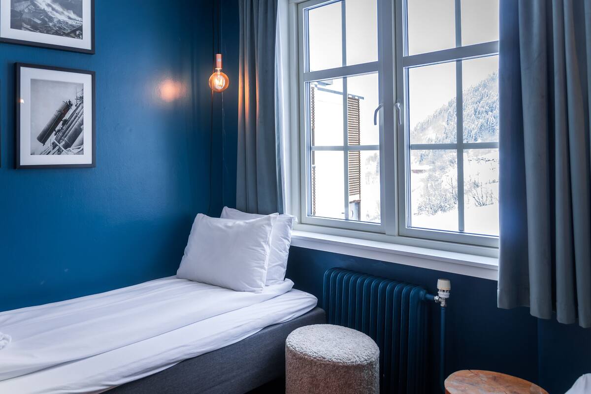 A room with a bed, a window with curtains, a radiator, and framed pictures on the wall.