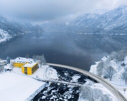 A snow-covered landscape with a yellow building, a bridge, and a lake surrounded by mountains.
